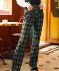 A person standing in a cafe wearing high-waisted 70's inspired green plaid wide-leg trousers with pockets.