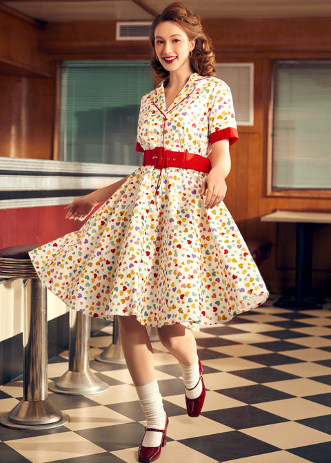 A woman standing in a vintage-style diner wearing a cream-colored belt with red accents and a red and multicolored print shirtdress.